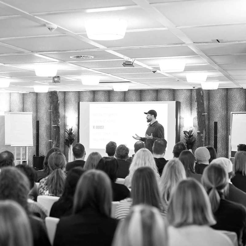 Das Bild zeigt eine Workshop Situation, in der man sitzende Personen von hinten erkennt die einem Mann zuhören, der etwas präsentiert. 