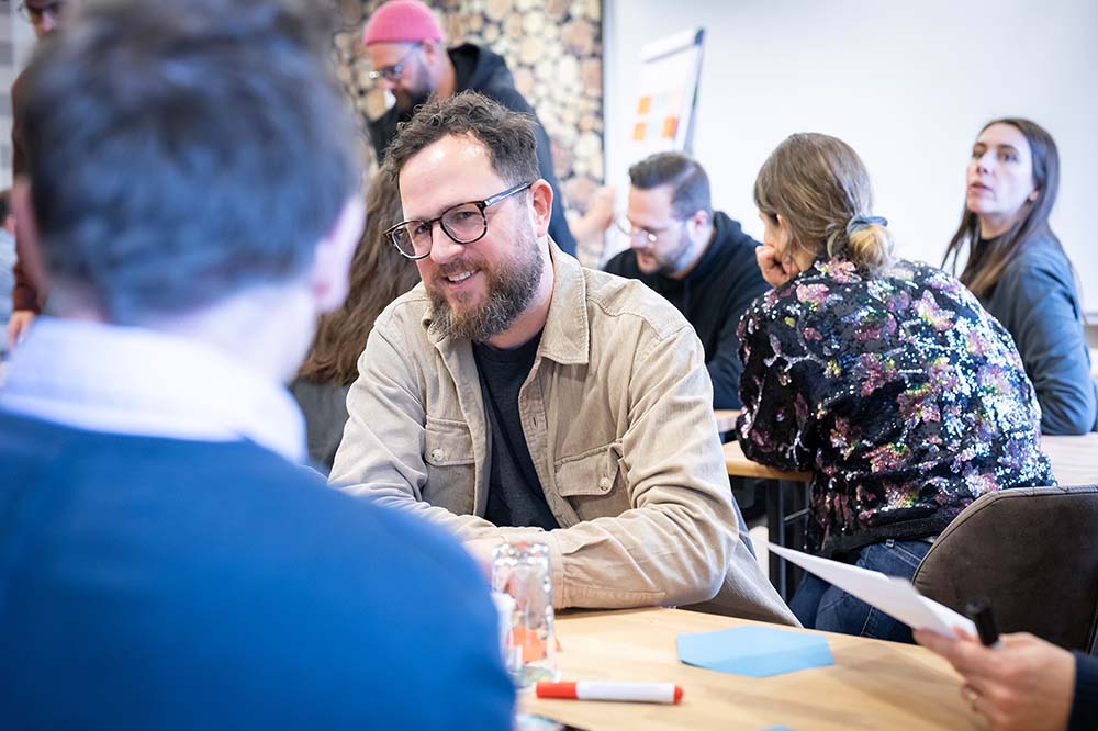 Mehrere Mitarbeiter werden in einer Meeting-Situation an Tischen sitzend gezeigt, während der Fokus auf einem lächelnden Mann mit Brille liegt.  