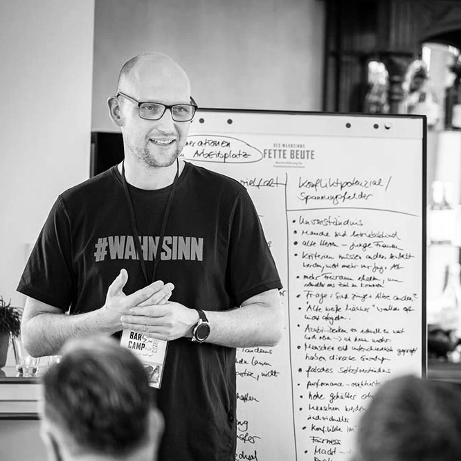 Ein Mann mit Brille und T-Shirt steht vor einer Tafel mit Notizen. Er spricht mit einer Gruppe von Personen und gestikuliert, während er seine Gedanken teilt. Die Atmosphäre wirkt engagiert und informativ.