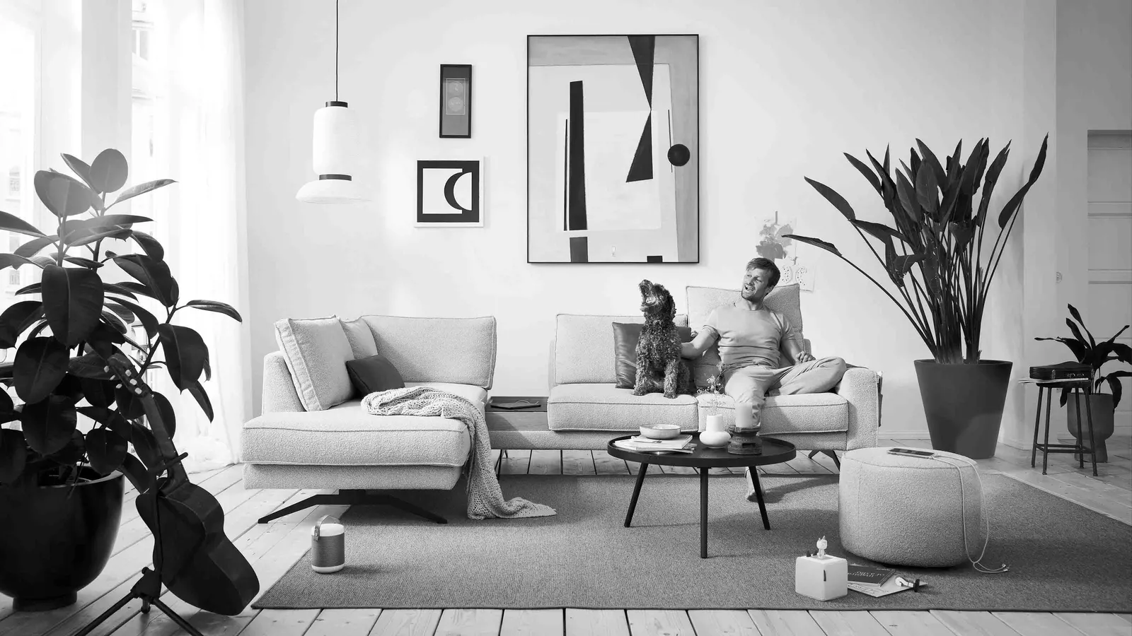 Abgebildet ist ein Wohnzimmer mit einem Mann, der mit seinem Hund auf einem Sofa sitzt.