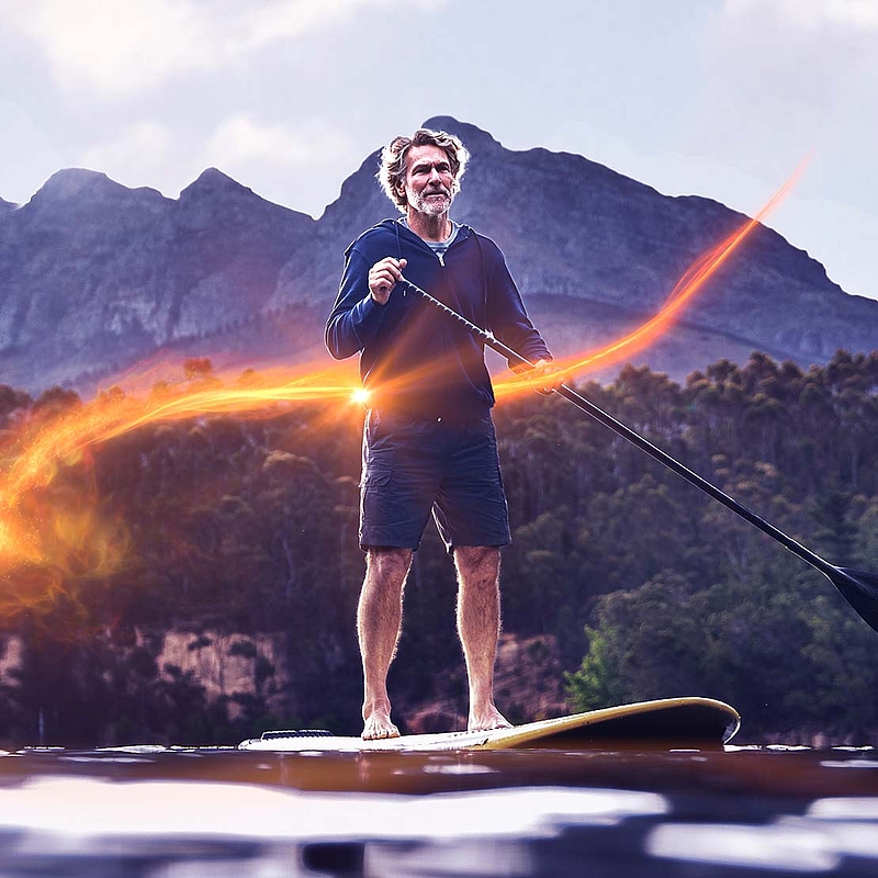 Ein älterer Mann steht auf einem Steh-Paddleboard auf einem See mit Bergen und Bäumen im Hintergrund.