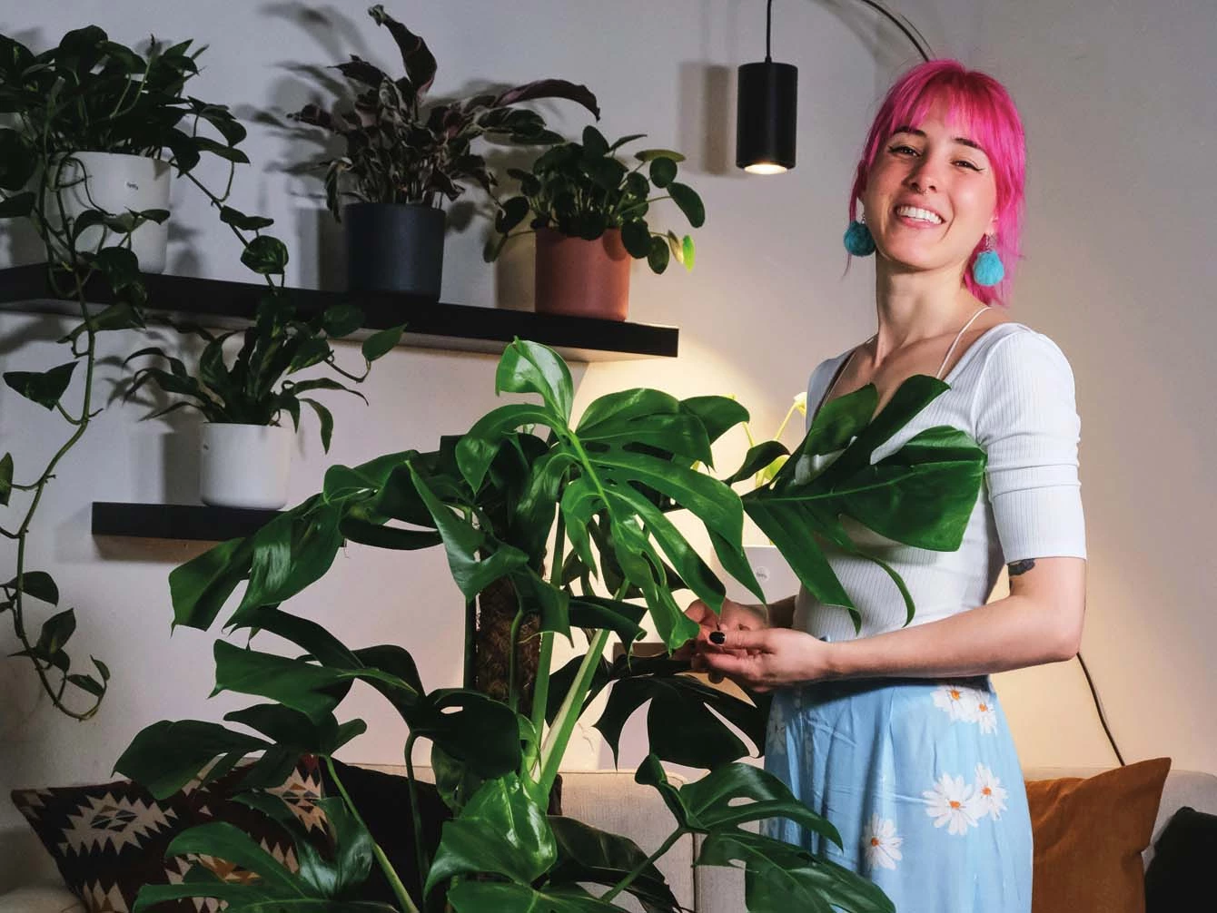 Eine Frau mit pinken Haaren und großen Ohrringen steht in einem Zimmer mit grünen Zimmerpflanzen und lächelt in die Kamera.